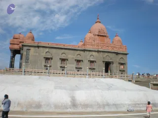 A photograph of a large grey stone temple-style memorial with red-orange domes, pillars at its left end and small barred windows along the wall, standing on a raised white stone platform under a blue sky with scattered clouds. Two people stand in the foreground at the platform's base: a man in a light grey shirt and dark trousers and a child in a pink top. Several small figures can be seen on the upper walkway at the right.