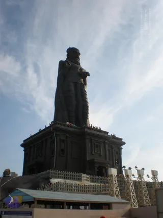 Low-angle photograph of a very tall dark grey statue of a standing male figure in dhoti-like robes with one hand raised near the chest, set atop a large stepped stone plinth against a pale blue sky with wispy clouds. Small distant people stand on the plinth balcony and along the railings, and floodlight towers flank the base. A tin-roofed shelter and a fenced walkway are in the foreground.