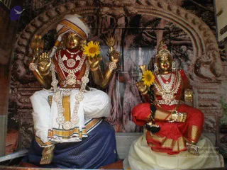 Two golden deity murtis stand side by side in a shrine against a backdrop of carved stone arches. The figure on the left, a four-armed deity murti with a white turban, wears a white dhoti with a blue border, layered white and red jewelled necklaces, and holds a yellow sunflower in one hand. The figure on the right wears a red sari with gold border, a jewelled crown and necklaces, and holds a yellow sunflower and a golden staff. Both are decorated with garlands and ornaments, with the figures arranged as a pair in a temple-style setting.