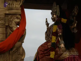 A photograph of a golden deity murti seated on an ornate golden ratha or palanquin canopy, with a red cloth draped over a carved golden pillar at left. The murti wears a tall crown, a red and maroon silk garment, garlands of yellow marigolds and dark red flowers, and holds a small bird-topped staff in one hand. Behind it, an overcast grey sky and distant hills are visible through the open side of the canopy.