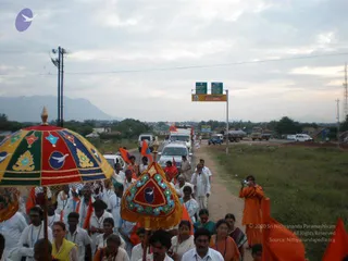 A photograph of a pada yatra procession on a rural road at dusk. Several people in white clothes walk behind two ornate decorated umbrellas or canopies, one multicoloured with a golden finial and a red-and-green mandala-like pattern, the other smaller and teal-and-red with a blue emblem. Many marchers carry saffron and orange flags, and a few women wear saffron and orange saris. A person in orange robes stands at right taking a photograph. A white SUV and other vehicles travel along the road beyond the crowd, with a green road sign and a distant hill range behind. Overall the scene is a roadside procession of devotees.