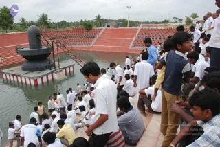 Outdoor photo of a large rectangular pool with a tall black stone Shivling on a square platform at its left edge, rising from a tiered base with a red-and-white striped plinth. Many men and boys in white shirts and white dhotis or trousers stand and sit along the stone steps and in the shallow water, some bathing or wading. On the right, a young boy in a blue striped shirt and khaki trousers stands watching, and a man in a blue shirt stands near the steps. Red-tiered stands and a palm-lined grounds stretch behind under an overcast sky.