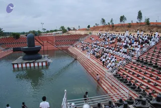Outdoor photograph of a large stepped tank with still water beside a massive black stone Shivalingam mounted on a square white-and-red base in the water. Rows of red stone steps on the right hold a crowd of young people seated in white clothing, many in white shirts and trousers, with a few in darker outfits. Round black stone pedestals line the lower steps. A small child in red stands at the water's edge, and a few people in white stand at the lower left and near the railing. A grey overcast sky, palm trees, and an orange boundary wall fill the background.