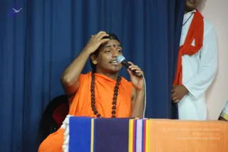 A photograph of a young man in orange saffron robes seated behind a draped table covered with a blue, purple and orange striped cloth. He holds a microphone to his mouth with one hand while the other hand rests on his head. He wears a rudraksha mala of dark beads and a red mark on his forehead. Behind him stands a second man in white clothes with an orange-red shawl over his shoulder, and a dark blue curtain hangs in the background. The scene appears to be a speaking event or youth gathering.