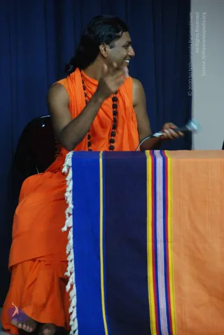 Photo of a man seated on a dark chair behind a table draped with a blue, navy and orange striped cloth with white fringe. He wears an orange sleeveless garment with an orange wrap and a long rudraksha mala. He has long dark hair swept back, smiles and gestures with one raised hand while holding a small white object in the other. Behind him hangs a deep blue curtain.