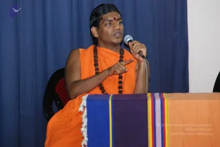 A man seated at a draped table speaks into a handheld microphone while pointing with his right index finger. He wears a sleeveless saffron-orange garment and a long rudraksha mala of dark brown beads, with a red mark on his forehead. He sits in a black chair before a dark blue curtain. The table is covered with a cloth in blue, purple, yellow and orange stripes with white fringe.