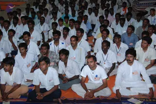 A large seated audience of young men fills the frame, sitting cross-legged on colourful striped woven mats and orange cloth. Most wear white shirts and white or cream trousers, many with lanyard ID badges and name cards. Some wear blue or striped shirts, and one man in the foreground wears a white shirt with a blue circular logo and a pendant necklace. Attention is directed forward toward an unseen speaker, with several men sitting attentively with hands folded. The setting appears to be a hall with a pale wall, and a partial white banner with red lettering is visible at the bottom right edge.