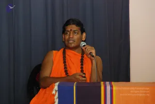 A photograph of a man in an orange saffron robe with a dark rudraksha mala around his neck and a small red bindi on his forehead, seated and speaking into a handheld microphone with his other hand resting on his lap. Behind him is a dark blue curtain and a pale wall, and in front of him is a table draped with a striped blue, purple, red and orange cloth. The setting resembles a seated talk or youth meeting.