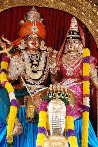 A photograph of a pair of decorated metal murti statues of a deity couple, the male figure on the left holding a trident and the female figure on the right in a pink and red sari with a crown. Both are adorned with jewelled necklaces, earrings and yellow and purple flower garlands. The male figure wears an orange turban and a dark jewelled garment, and a silver kalasha-like vessel stands in front of them. A red curtain and gold-framed arch form the backdrop. Statues only; no living people are present.
