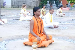 A photo of a group meditation outdoors on a dusty ground with a small pile of grey ash or stone chips. In the foreground a man with long dark hair tied back sits cross-legged in saffron-orange robes, wearing a rudraksha mala and a red tilak on his forehead, eyes closed, hands resting on his knees. Beside him is a brass kendi or oil vessel. Behind him several people in white clothes with yellow shawls sit in meditation, and a row of stone statues and a red signboard appear in the background beneath thatched fencing.