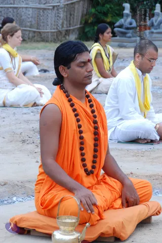 A photograph of a seated meditation gathering on bare sandy ground. In the foreground a man with long dark hair tied back sits cross-legged on an orange cushion, eyes closed, hands resting on his knees. He wears an orange sleeveless robe and a long dark rudraksha mala, with a red bindi on his forehead. A brass vessel with a spout sits in front of him. Behind him sit other meditators: a woman in white with a yellow shawl, a woman in white and yellow, and a bespectacled man in white clothes with a yellow shawl, all with eyes closed. Stone sculptures of seated figures and a thatched bamboo fence stand in the background.