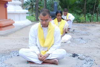 Three devotees sit cross-legged outdoors on sandy ground with ash scattered around them, appearing to be in a fire-related ritual setting. In the foreground a shaven-headed man in white clothes and a yellow shawl sits with his hands joined in prayer, wearing glasses and a red tilak on his forehead. Behind him a woman in white with a yellow shawl and a red bindi sits with her eyes closed, and further back a young man in white with a yellow shawl sits with his hands folded. A white and orange stone structure and palm trees stand in the background.