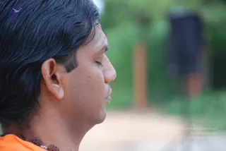 Close side profile photograph of a man with eyes closed, dark hair swept back, wearing an orange garment and a dark rudraksha mala at the neck. The background is a blurred green garden with a brown wooden post and a dark-clothed figure far behind. The man appears to be in a calm, meditative or contemplative moment outdoors.