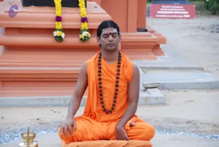 A man seated cross-legged in meditation on a sandy ground in front of an orange-painted temple structure. He wears an orange saffron-coloured sleeveless garment with a matching shawl draped over one shoulder, a long rudraksha mala of dark beads around his neck, and a red bindi on his forehead. His eyes are closed and his hands rest on his knees. Behind him, yellow and white marigold garlands hang from the orange plinth, and a red board with white lettering sits on the right. A brass oil lamp and a small brass dish of ash are placed in the foreground.