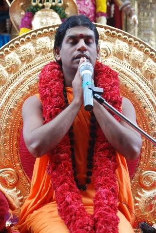 A seated man in an orange saffron robe sits on an ornate gilded throne with floral and scrollwork carving and a round halo-like back. He wears a thick red flower garland and multiple dark rudraksha beads, with a red tilak on his forehead. His eyes are closed and his palms are pressed together in namaste in front of a silver microphone on a boom stand. Behind him are pink and yellow flower garlands and a brass kalasha. a person is seated in devotional posture, consistent with the saffron robe, rudraksha malas and garland cues.
