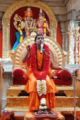 A man in orange saffron robes sits on an ornate gilded throne with red cushions, speaking into a microphone on a stand. He wears a red flower garland, a white jasmine garland, and a rudraksha mala, with a red tilak on his forehead; his feet rest on a small cushion with flower petals. Behind him stands a gilded arch framing two golden deity murtis, one with an orange turban and trident-bearing hands and one in a pink sari with a crown, above a red curtain. Golden lamp stands flank the right side.