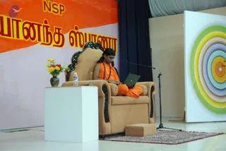 A photograph of a stage event. A man with long dark hair sits on a tan upholstered throne-style chair with black carved trim, wearing orange robes and a rudraksha mala, working on an open laptop balanced on his lap. A small stand with a water bottle and a vase of yellow and red flowers sits to the left, a microphone stand stands beside the chair, and a footstool rests on a patterned red rug. Behind him a large orange and pink banner carries the name NSP and Tamil script, and to the right a white board displays concentric colored circles in green, yellow, lilac and orange. No other people are visible.