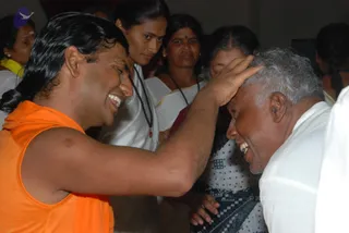 A man with long, swept-back black hair wearing a sleeveless saffron-orange top and an orange shawl places his right hand on the head of an older, grey-haired man in a white shirt, who smiles and bows his head. Behind them several women in white clothing stand watching, one wearing a dark beaded mala necklace and another in a white and teal patterned sari. The setting is an indoor room with plain walls. The orange-clad man has distinctive long hair and saffron clothing with a shawl, and the scene shows a blessing gesture; the page does not indicate his presence, so this appearance-based label is probable only. a person is the man in orange, giving a blessing to the older devotee.