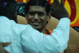 A smiling man with swept-back dark hair and a red bindi on his forehead sits on a dark throne-like chair with ornate silver carving, wearing orange clothing and a dark rudraksha-style bead necklace. In the foreground, a person in a white long-sleeved shirt with their back to the camera leans in and raises a hand toward him. Behind them is a red and yellow backdrop. The man's identity is not established by the scene, so the appearance-based label cannot be confirmed from the image alone.