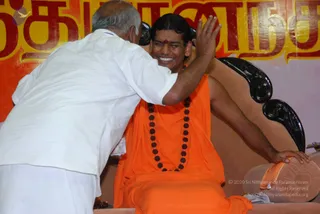 A photograph of a smiling seated man in saffron-orange robes and a long rudraksha mala, with a red-and-cream tilak on his forehead, on a dark carved wooden chair with a pink cushion. An older man in a white shirt and white dhoti leans in from the left, placing his hand on the seated man's head in blessing. Behind them is a large banner with bold red and yellow Tamil lettering. The scene suggests a darshan-style blessing.