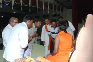 A photograph of a hall with maroon drapes and white fringed valances along the ceiling. In the right foreground, a figure with long dark hair and a sleeveless saffron-orange garment sits with their back to the camera on a pale peetha-style seat. Several men stand in front of them: on the left, a man in a white shirt and an older man in white with glasses press their palms together in anjali mudra; in the centre a man in a white dhoti and orange shawl stands beside a man in a striped navy-and-white polo. A tray of bananas and other offerings sits in the lower left.