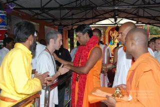 A photograph at an outdoor temple-style hall with a corrugated roof, where a man in an orange sleeveless robe and a long red flower garland stands at the centre, holding the hands of a shaven-headed man in white clothes and a rudraksha mala. Around them stand other men in white and saffron-orange robes; one man at left wears a yellow kurta, and a saffron-robed attendant at lower right holds a brass vessel. Behind them is a colourful deity murti in a shrine, with a bright orange and gold backdrop. Several people stand at the rail in the background.