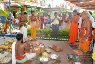 Outdoor temple ritual with a seated priest bare-chested with a white cloth over his shoulder, a red sacred thread and beaded necklaces, tending a fire pit of burning wood and offerings, with a brass bowl and a vessel of white sweets nearby. Around him are standing people: a man in a green and white dhoti holding a small brass pot, a man in a saffron dhoti with a folded hand gesture, a woman in saffron robes with a garland, and an older bald man in saffron dhoti and beaded malas with hands folded in prayer. Musicians with horns play nearby, a dhoti-clad man in white and others stand by. Behind them are colourful temple shrines, a painted lion statue, banana plants, marigold garlands and a canopy. Floral offerings and oil lamps are arranged on the left.