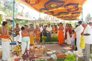 Outdoor temple ritual under a large orange canopy with red, green and yellow medallion patterns, supported by blue poles. A fire pit with burning flames sits on a paved platform in the centre, surrounded by brick-lined offering pots, flower-decked kalasha-style vessels and small brass bowls. A bare-chested priest in white dhoti with a red sash stands at the left, and a young man in orange robes with a red-and-orange shawl stands near the centre-right with hands folded. An older man in saffron-orange cloth and a rudraksha-style mala stands near the fire, and a man in white with a red-and-orange shawl and yellow bag stands at the right edge. A small girl in pink stands among the devotees. Behind them are multi-coloured temple structures, painted gopurams and a green deity statue with raised arms.