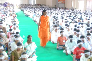 A large seated congregation of hundreds of people wearing white clothes and white blindfolds fills a hall, sitting on the floor on both sides of a long green carpet aisle. A single figure in flowing orange robes, with long dark hair pulled back, walks barefoot down the centre of the aisle. Among the seated people are a woman in a white sari with a teal border, a woman in a mustard-and-green sari, and a man in an orange shirt. Water bottles sit on the floor near the front. The photo is blurred, and the orange-robed walker is a person visible in the scene whose identity cannot be confirmed from appearance, so the a person field is unsure.