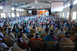 A large indoor hall filled with hundreds of seated devotees, most wearing white headbands and white or pastel clothing, including saris in blue, pink, orange and red. They sit in rows of yellow-and-white plastic chairs facing a stage. A tall orange-robed figure stands near the stage, and a large banner behind the stage shows a smiling figure in an orange turban beside a painted deity image. Two projection screens hang above the stage and white canopy fabric covers the ceiling.