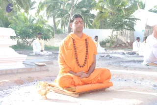 A photograph of a man seated cross-legged on an orange-covered wooden platform in an open outdoor yard with sandy ground and ash-grey patches. He wears an orange saffron robe, a long rudraksha mala with dark beads, and a red bindi on his forehead, with eyes closed and hands resting together in his lap. A small golden vessel sits on the ground beside him. Behind him, several men in white dhotis and yellow shawls sit in meditation, and palm trees and a white wall frame the scene. a person cues: orange saffron robe and rudraksha mala on the seated central figure.