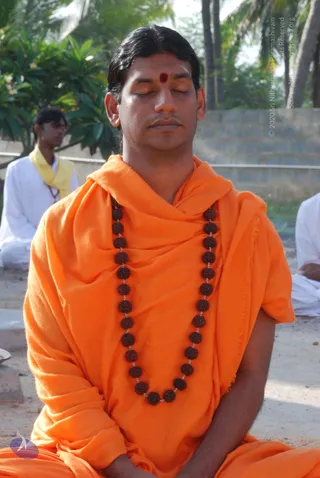 A photograph of a man seated cross-legged outdoors in meditation with eyes closed, hands resting in his lap. He wears an orange-saffron shawl draped over his shoulders and a long dark rudraksha mala with large beads around his neck, and has a red tilak mark on his forehead with swept-back dark hair. Behind him, a man in white clothing with a yellow scarf sits with eyes lowered, and palm trees, plants and a concrete wall form the background. An appearance-based a person candidate in orange with rudraksha mala is seated in meditation.