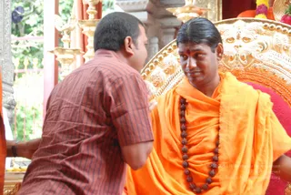 A seated man in saffron-orange robes and a long rudraksha mala sits on an ornate gold throne with a red cushion, a small red mark on his forehead. A devotee in a brown-and-red striped short-sleeved shirt leans in toward him from the left, one hand raised near the throne. Behind them are gold lamp stands and a gilded metal gate with green foliage beyond, and a faint decorative setting suggests a temple sarva darshan setting.