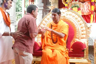 A photograph of a seated figure in an ornate gold throne with red cushions, wearing saffron-orange robes and a dark rudraksha mala, with hair swept back, extending hands toward a man in a red-and-brown striped short-sleeved shirt and khaki trousers who bows forward with palms together. Behind the throne stands a deity murti draped in red and orange cloth, adorned with yellow and pink flower garlands, with a brass oil lamp. A man in white clothing with an orange shawl stands at the left edge. The setting is a temple-like pavilion with carved stone columns and greenery beyond.