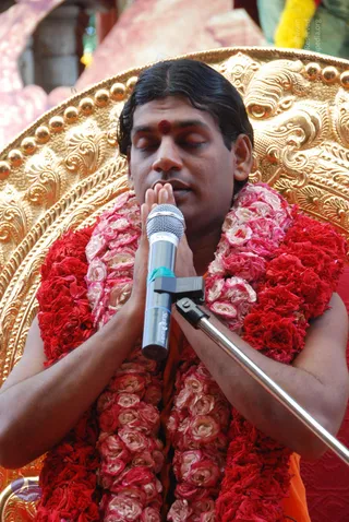 A man with swept-back dark hair and a red tilak on his forehead sits with eyes closed, palms pressed together in namaskar before a microphone on a stand. He wears a garland of red and pink roses and an orange saffron garment. Behind him is an ornate gold-embossed throne with a rounded backrest.