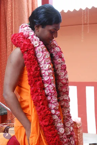 A man in an orange saffron robe stands in profile with his head bowed, wearing several long garlands of red and pink-white roses draped over his shoulders and chest. Behind him are orange patterned curtains and a red and white striped wall, with a small wooden bead mala visible at the lower left. The garlands dominate the frame and suggest a felicitation or darshan setting.