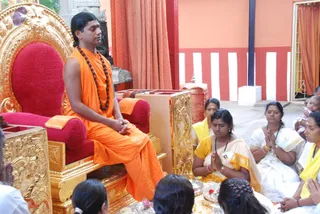A man in orange saffron robes and a rudraksha mala sits cross-legged on an ornate gold-and-red throne with his hands folded, eyes closed, in a courtyard with red and white striped walls and orange curtains. A row of devotees sits on the floor before him with palms pressed together in namaskar: a woman in a yellow-bordered sari with a name badge, a woman in a white sari with gold bangles, and a woman in a yellow sari beside them holding a baby. Brass plates with flowers and offerings lie in front of the throne. The man is a probable Swamiji on the throne, shown with long swept-back hair, saffron robes and a rudraksha mala.