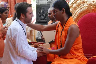 A photograph of a man in a white collarless kurta with a dark rudraksha mala kneeling close to a seated figure in an orange saffron garment and a long dark mala. The seated figure has long dark hair swept back, sits on a red cushioned throne with ornate gold carved backrest, and holds the kneeling man's hands. Behind them a bald man in orange and a woman in orange and yellow stand near a black stone deity murti of Ganesha with a small lamp. A wooden box with red cord and rudraksha beads sits in front. The seated figure matches the Swamiji (probable) description through long swept-back hair, saffron clothing and rudraksha mala, seated on a throne in a devotional darshan setting.