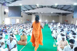 Photograph from behind of a large hall filled with seated participants in white clothing and white blindfolds. In the foreground, a figure with long dark hair wears a flowing saffron-orange garment and raises one hand toward the crowd, walking along a central green carpeted aisle. Rows of seated devotees in white and some in colourful saris and shirts fill both sides. White ceiling canopies, ceiling fans, dark projection screens, and a water bottle on the floor are visible. Overall, a dense group session in progress.