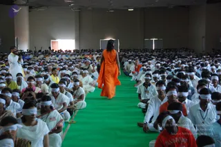 A large hall filled with hundreds of seated participants wearing white clothes and white blindfolds over their eyes, sitting in rows on both sides of a green carpeted central aisle. A single figure in a long flowing orange robe with long dark hair walks away from the camera down the green aisle, toward the far end of the room. A cameraman in white clothing films at the left edge. Some participants wear red or orange shirts, and one person in the foreground wears a red shirt printed with the word PLAYSPOINT. Plain beige walls and tall windows frame the space.