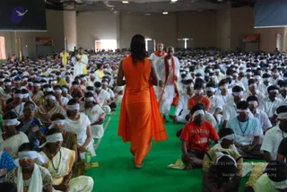 A large indoor hall full of seated participants, most wearing white head blindfolds, with a long green carpet running down the center. In the foreground a figure in a flowing orange robe with a long orange shawl walks away from the camera along the carpet, barefoot, with long dark hair. Behind and to the side, a bald man in white clothes with an orange shawl walks alongside. Many participants wear white, some in saffron and yellow; a woman in a yellow-and-white sari sits at left. Volunteers in white and yellow stand near the back. Banners hang on the walls and a water bottle sits on the carpet edge.
