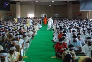 A large hall filled with hundreds of seated devotees wearing white clothes and white blindfolds over their eyes, arranged in rows on both sides of a central green carpet aisle. Near the front, a man in white robes walks along the aisle, and a woman in a bright orange sari walks behind him toward the far end. Other attendees in white and in colourful saris and shirts sit in the rows, with a few orange-clad figures visible among them. Overhead are ceiling fans, white walls, and banners on the side walls. The scene suggests a guided blindfolded meditation session in a large ashram hall.