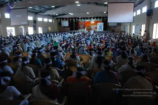 A large hall filled with many seated devotees, most wearing white headbands and white or light clothing, seated in rows of plastic chairs facing a stage at the far end. On the stage backdrop is a large banner portrait of a saffron-robed figure with a turban and a smiling face, alongside a decorative poster; a small figure in white stands near the stage. Two large projector screens hang from the ceiling, and white draped fabric and ceiling lights are visible above.