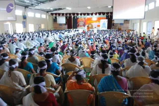 A large hall packed with seated devotees, most wearing white headbands, sitting in rows of cane-backed chairs facing a stage. Many women wear white, cream, blue, teal and orange saris and salwar outfits. At the back of the stage hangs a large framed portrait of a smiling man in an orange turban, with a Tamil banner beside it and a second framed image of a figure in blue. Two ceiling-mounted projection screens and white drapes frame the hall. Several people in white stand near the stage and aisles.