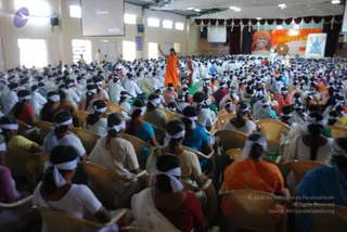 A large hall packed with hundreds of seated participants, mostly women, wearing white headbands and white or light-coloured clothing, sitting in rows of cane chairs. In the foreground a figure in a bright orange robe stands on a green carpeted aisle with an arm raised, while another figure in white walks beside them. At the back, a stage displays a large orange-and-red banner with a portrait of a smiling man in an orange turban and saffron robe, and a smaller framed picture of a seated deity-like figure in blue. A projection screen and white ceiling lights hang above the hall, with white curtains framing the stage.