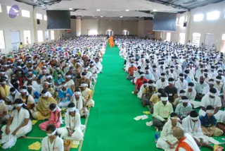 A photograph of a large hall with a long green carpet running down the centre aisle between rows of seated devotees. Hundreds of participants sit cross-legged on the floor wearing white headbands over their eyes, many in white or light clothing, with some in saffron, pink, green and blue. Small yellow sheets lie on the carpet near the seated rows. At the far end of the aisle a single figure in an orange-saffron robe stands facing the crowd. Large dark screens hang from the white ceiling, and tall windows line the walls.