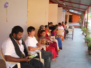 A covered veranda with red-painted steel roof posts and potted plants lines the outside of an orange-walled building. Several people sit in beige plastic armchairs along the wall: a man in a white shirt with a dark grey shawl and beaded necklace holds a small green packet, beside a woman in a white t-shirt and grey trousers, then a boy in an orange-and-black striped shirt, and women in red, orange and blue saris. Further back, a row of small bottles sits on a metal side table, and a hanging sign is visible on the wall. No a person candidate is visible.