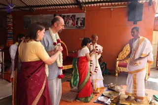 A ceremonial exchange in an orange-walled hall with a corrugated roof. A man in a grey shirt and grey trousers and a woman in a mustard top and maroon sari stand at left, both wearing white and red flower garlands, holding hands. Beside them a young girl in a red and green sari and a bald man in white dhoti and shawl with a flower garland stand before a brass tray and small bowls on a mat. At right a young man in a white dhoti with gold border, bare-chested, wears beaded necklaces and gestures toward the tray. Gold-framed chairs with red cushions sit along the wall, and a framed photo display hangs above.