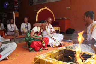 A homa fire ceremony on an orange floor mat. In the centre, a bald man in white dhoti and shawl with white flower garland and red floral garland sits cross-legged beside a woman in a red and green sari with flower garlands, her hands pressed together as he holds her head. Behind them a red throne with gold trim stands against an orange wall. To the right a priest in white with a gold chain and rudraksha-style beads tends the fire. Seated devotees in white and grey clothing line the background, and a fire pit with flames and marigold petals lies in the foreground.
