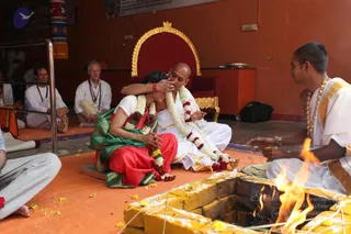 A photograph of a homa at an ashram hall with orange walls and a golden-framed red throne behind. A bald man in white ritual clothes with white and red flower garlands sits on a mat, while a woman in a red and green silk sari with bangles and red-and-white garlands leans her head against his chest and is embraced by him. A young priest in white with a gold-bordered shawl and beaded necklace sits at right, tending a fire in a yellow brick fire pit with flames and scattered flower petals. Seated devotees in white clothes, some wearing rudraksha malas, watch from the left, and a metal railing stands nearby. The figure in white with the garlands is a probable match for a person in the appearance-based label.